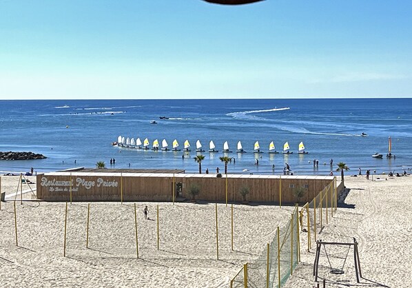 Une plage bordée d’une rangée de voiliers blancs, d’une promenade en bois et d’un panneau indiquant le restaurant Plage Privée.
