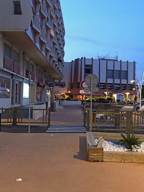 Une vue de rue avec un bâtiment moderne doté d’une façade originale, un passage piéton et un petit espace jardin agrémenté de jardinières en bois.