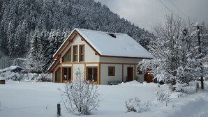 Una casa de dos pisos amb un teulat cobert de neu i façana de fusta, envoltada d'arbres plens de neu i una muntanya al fons.