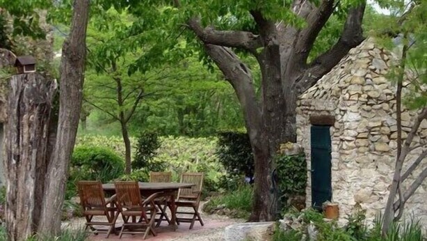 A stone building with a green door, surrounded by trees and a gravel path leading to a wooden table and chairs.