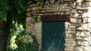 A rustic stone building with a green shuttered window, surrounded by lush greenery and purple flowers.