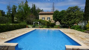 A rectangular swimming pool with a stone border, surrounded by a garden with various plants and a house in the background.