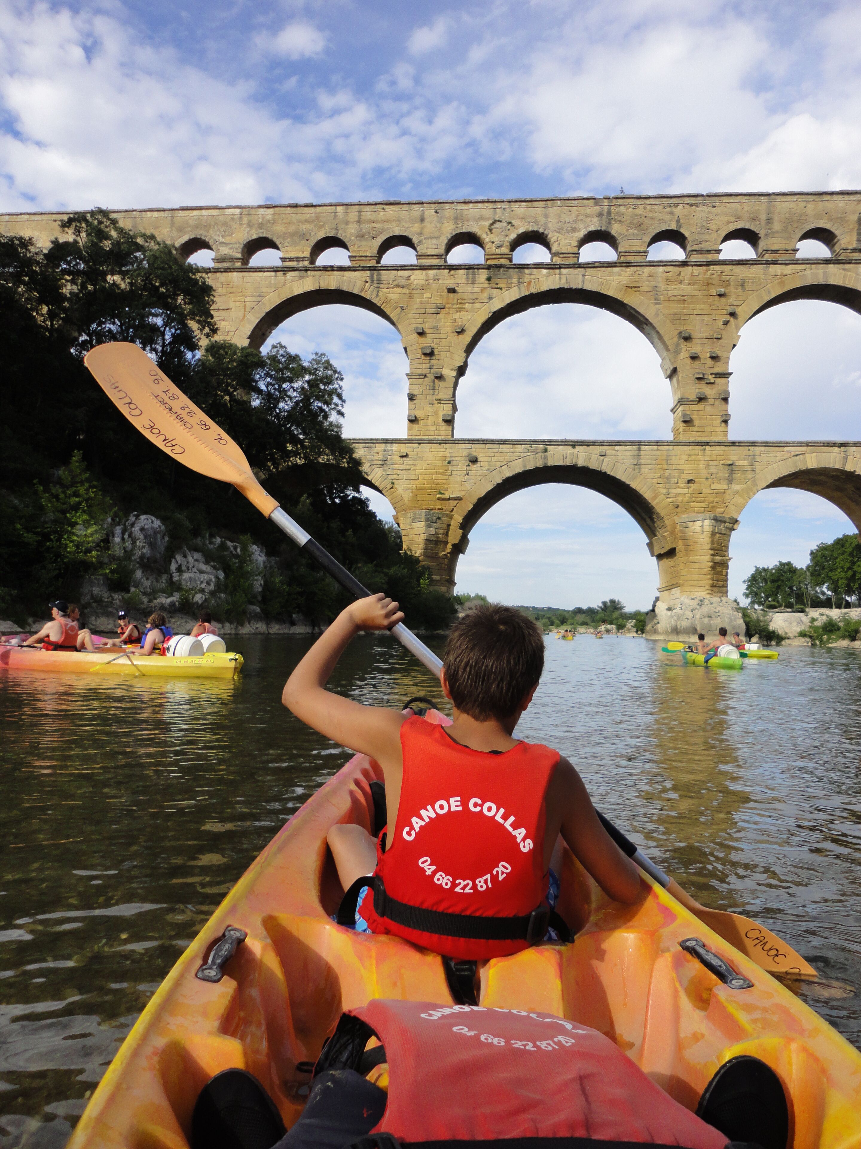 A person in a canoe with a life jacket labeled "Canoe Collas" paddling under a stone bridge.