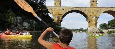 A person in a canoe with a life jacket labeled "Canoe Collas" paddling under a stone bridge.