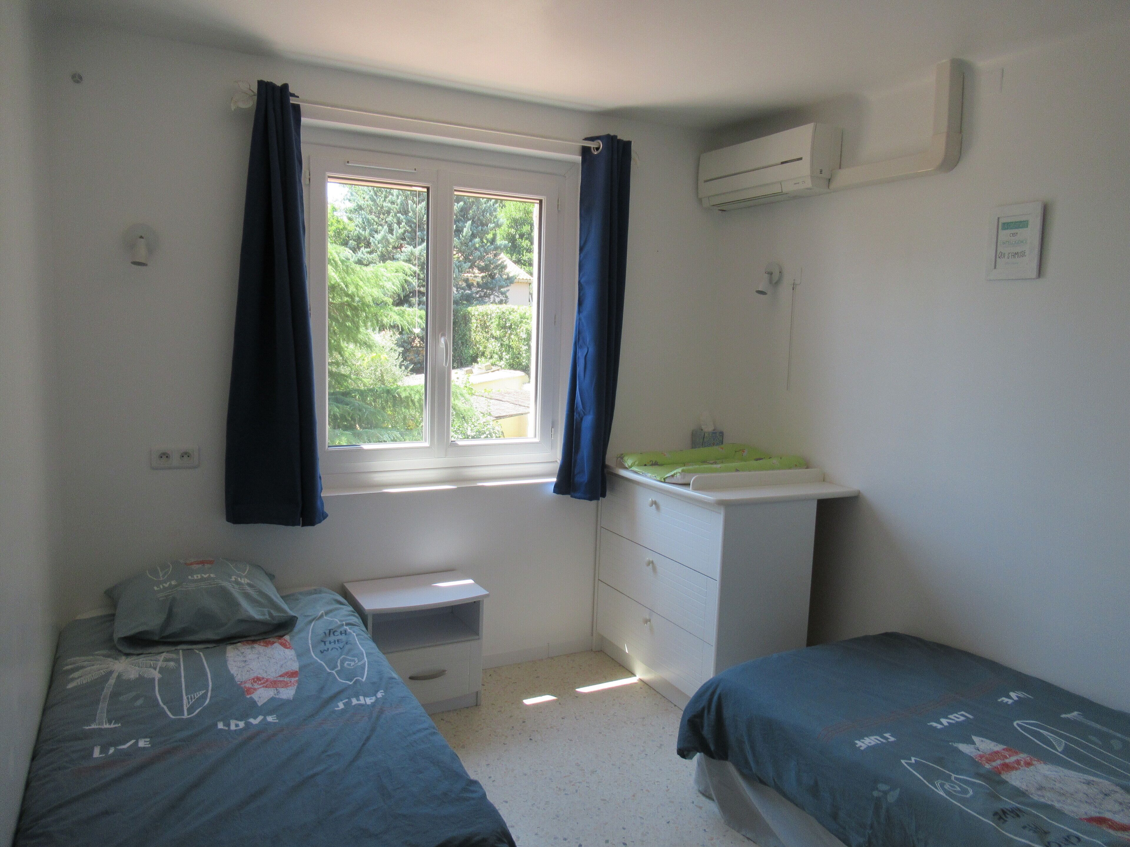 Un dormitorio con dos camas individuales, una ventana con cortinas, un aparador blanco y un equipo de aire acondicionado.