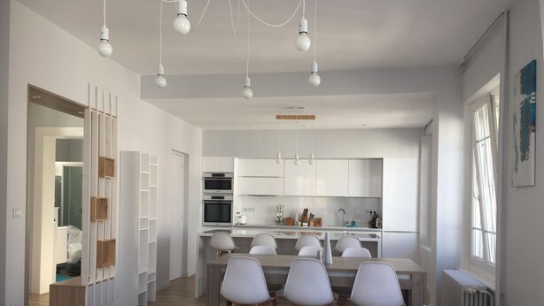 Una cocina moderna con armarios blancos, un comedor con sillas blancas y una sala de estar con sofá blanco y cojín azul.
