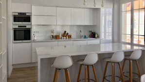 Una cocina moderna con armarios blancos, una isla central con taburetes altos, hornos integrados y un amplio ventanal que proporciona luz natural.