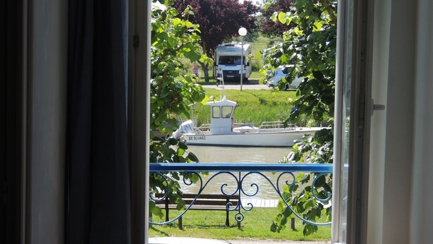 Blick durch ein Fenster mit blauer Geländer, ein Boot und viel Grün draußen.