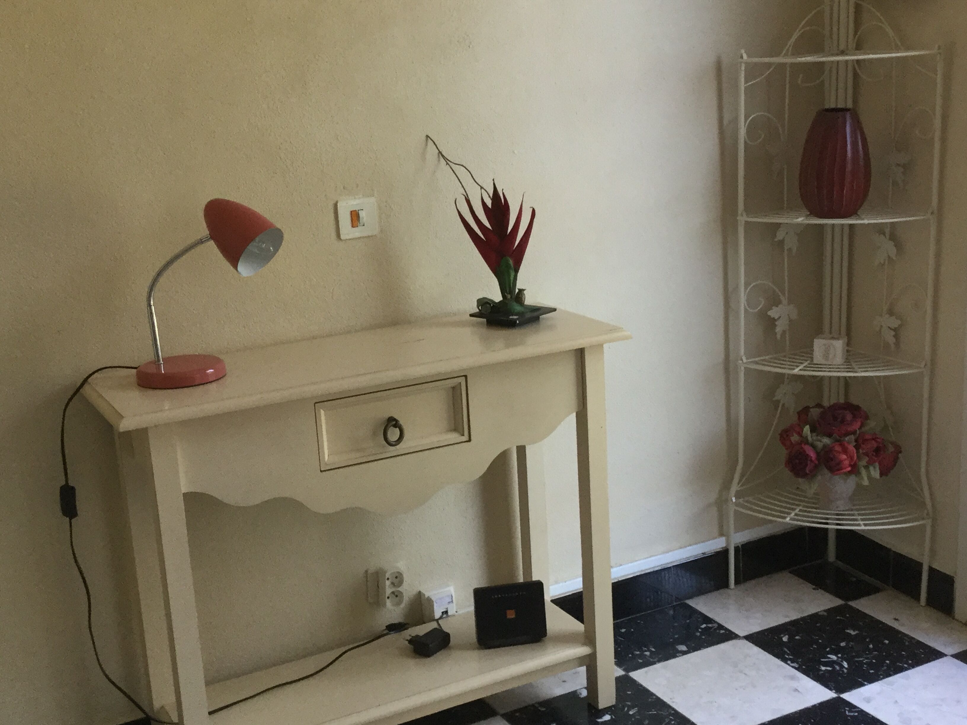 A desk with a lamp, a small plant, and a laptop. There is a power outlet and a shelf with a vase and flowers.