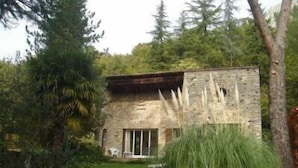 Ein steinernes Gebäude mit geneigtem Dach, umgeben von üppiger Vegetation und hohen Bäumen.