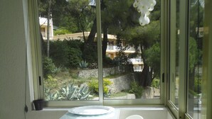 A small room with a table and four chairs, a window with a view of greenery and buildings, and a hanging light fixture. - A beautiful, quiet 3-star apartment in a wooded park with a view of the bay (Cassis)