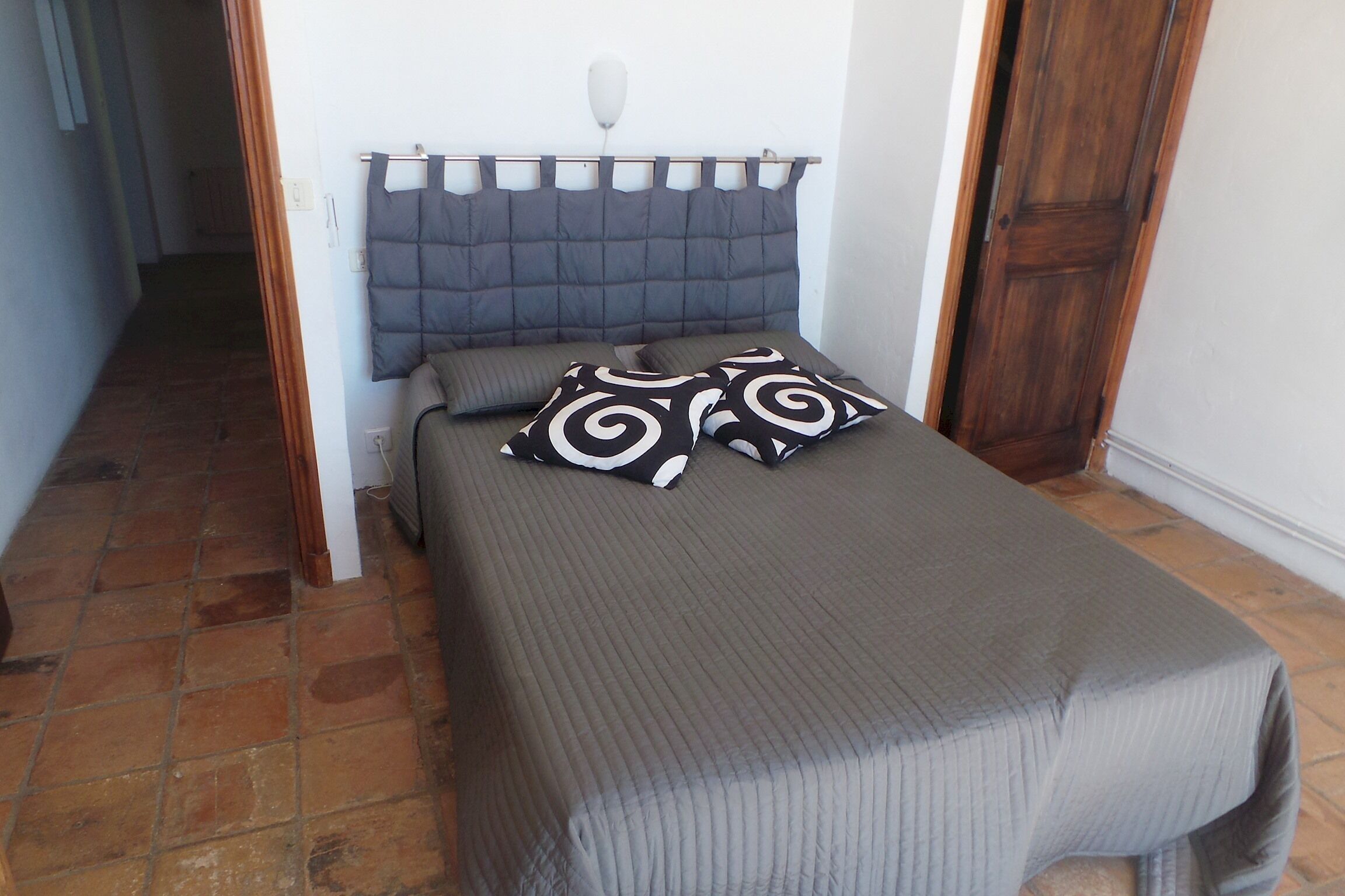 Een klein, netjes opgemaakt bed met een grijze spreidek en twee gestreepte kussens, een houten hoofdbord en een betegelde vloer.
