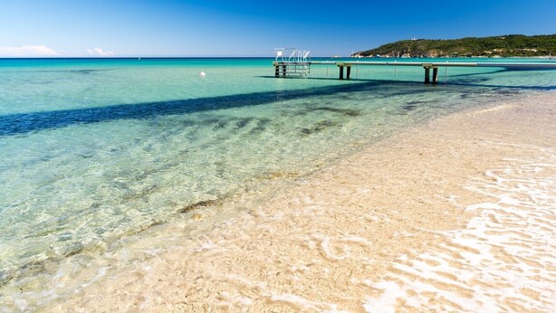 Una playa despejada con un muelle que se adentra en aguas turquesas, una orilla arenosa y una colina con árboles al fondo.