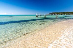 Ein klarer Strand mit einer Pier, die sich ins türkisblaue Wasser erstreckt, ein sandiger Uferstreifen und ein Hügel mit Bäumen im Hintergrund.