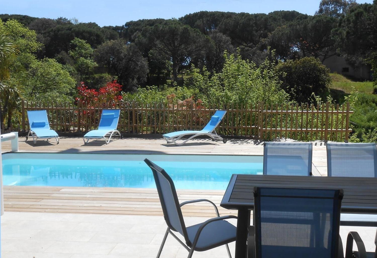 Zona de piscina amb gandules i una taula, envoltada per un pont de fusta i una tanca.