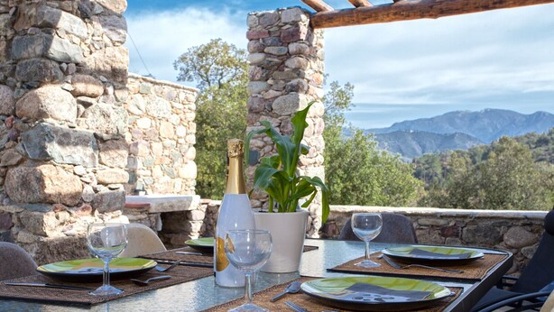 An outdoor dining area with stone walls, a table set for two with wine glasses and plates, and a view of mountains in the background. - MONTE-CINTO Stone house -PAGLIAGHJU- Gorges de l'Asco, not far from the sea  (Moltifao)