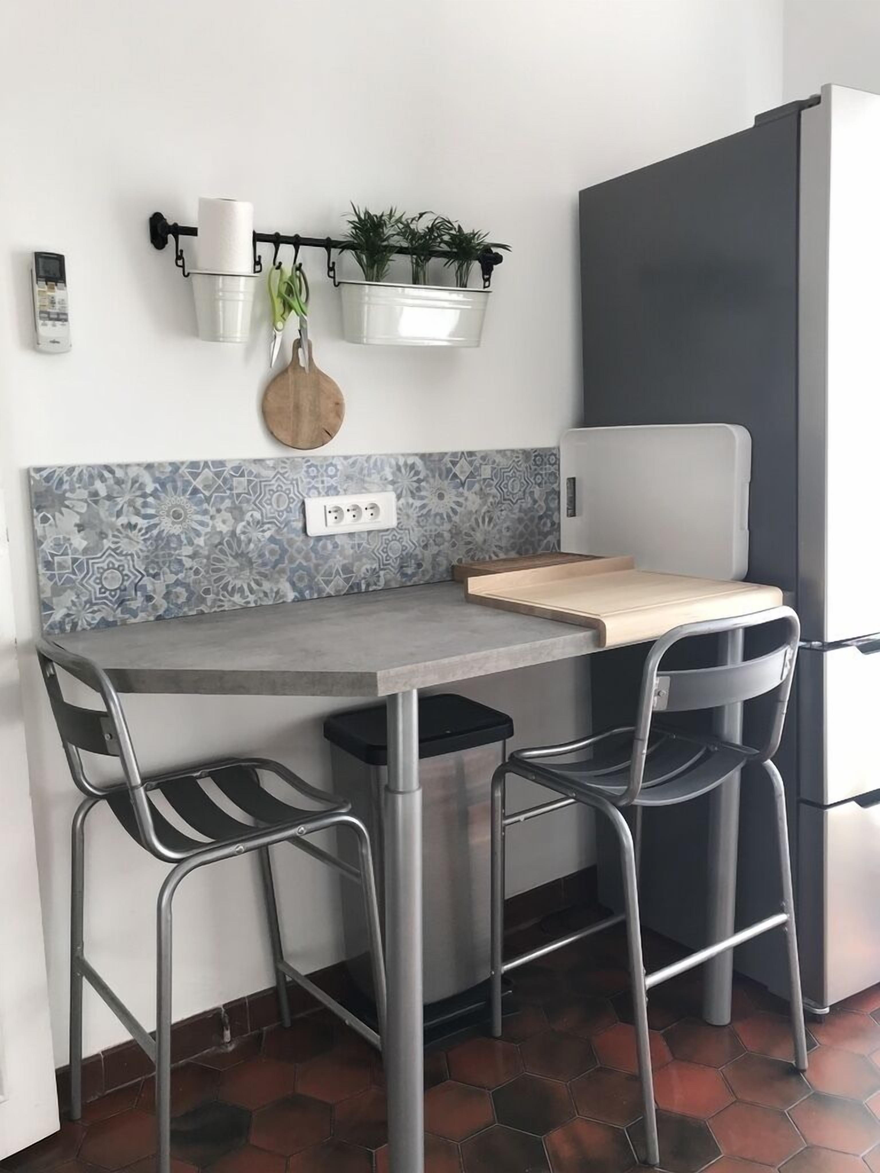 Eine kompakte Küche mit grauer Arbeitsplatte, zwei Metallstühlen, einem kleinen Holztisch, einem weißen Kühlschrank und einem an der Wand befestigten Pflanzenhalter.