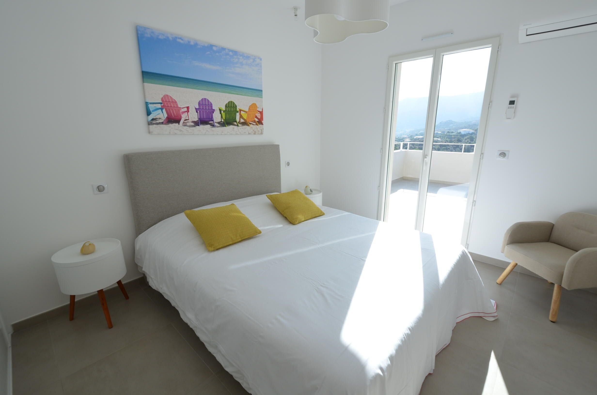 A bedroom with a bed, a chair, a painting of beach chairs, and a balcony with a view.