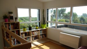 Un coin lecture avec une bibliothèque en bois, un fauteuil violet et une vue sur la verdure grâce à de grandes fenêtres.