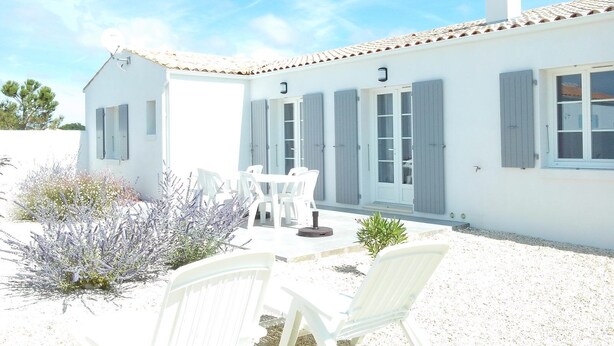 A white building with a tiled roof, a chimney, and a covered patio area with white chairs and a small table.