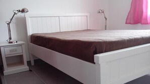 A bedroom with a white bed, a nightstand, a lamp, and a window with a pink curtain.