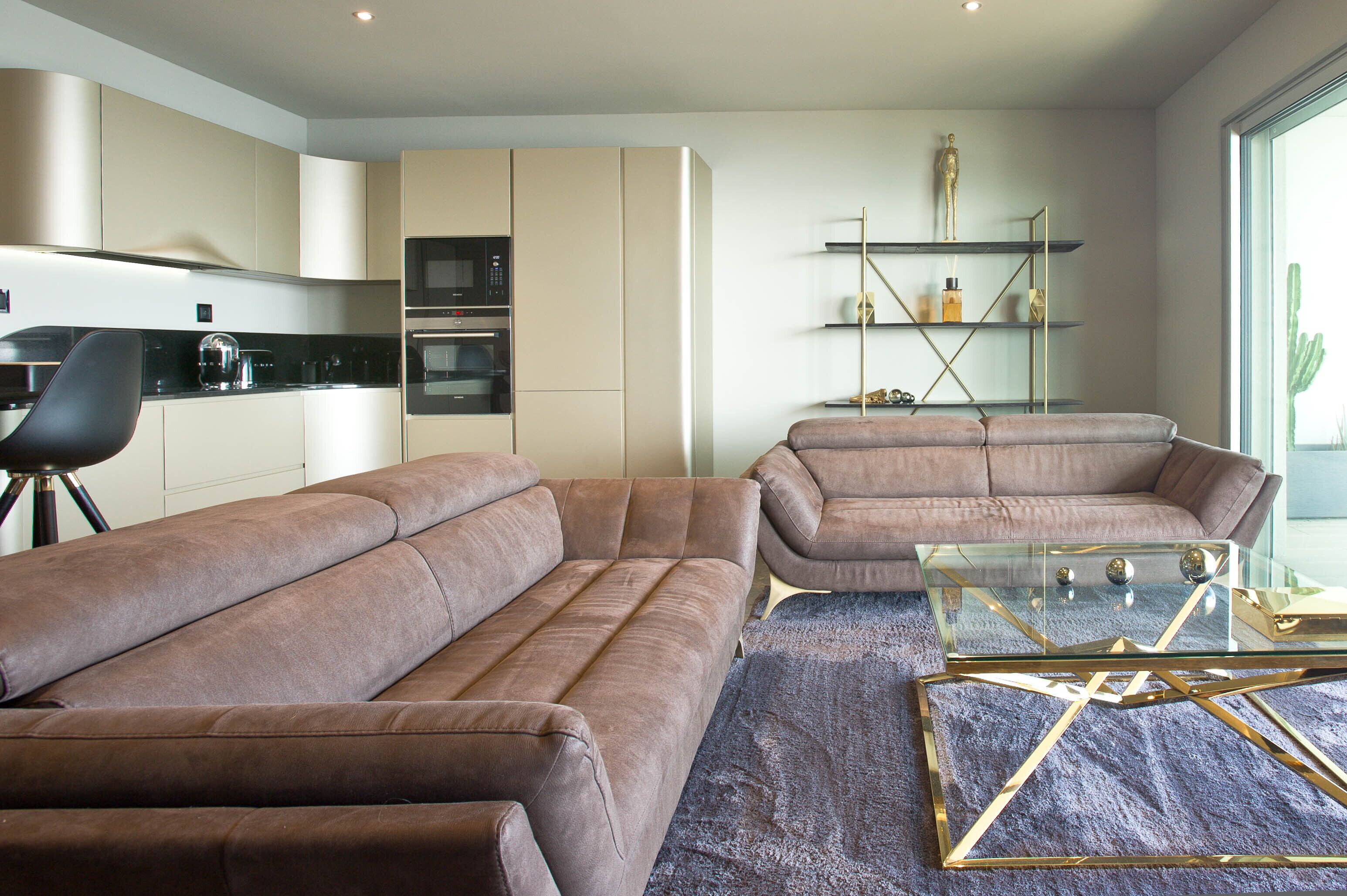 A modern living room with a sectional sofa, a glass coffee table, and a kitchen area with built-in appliances.