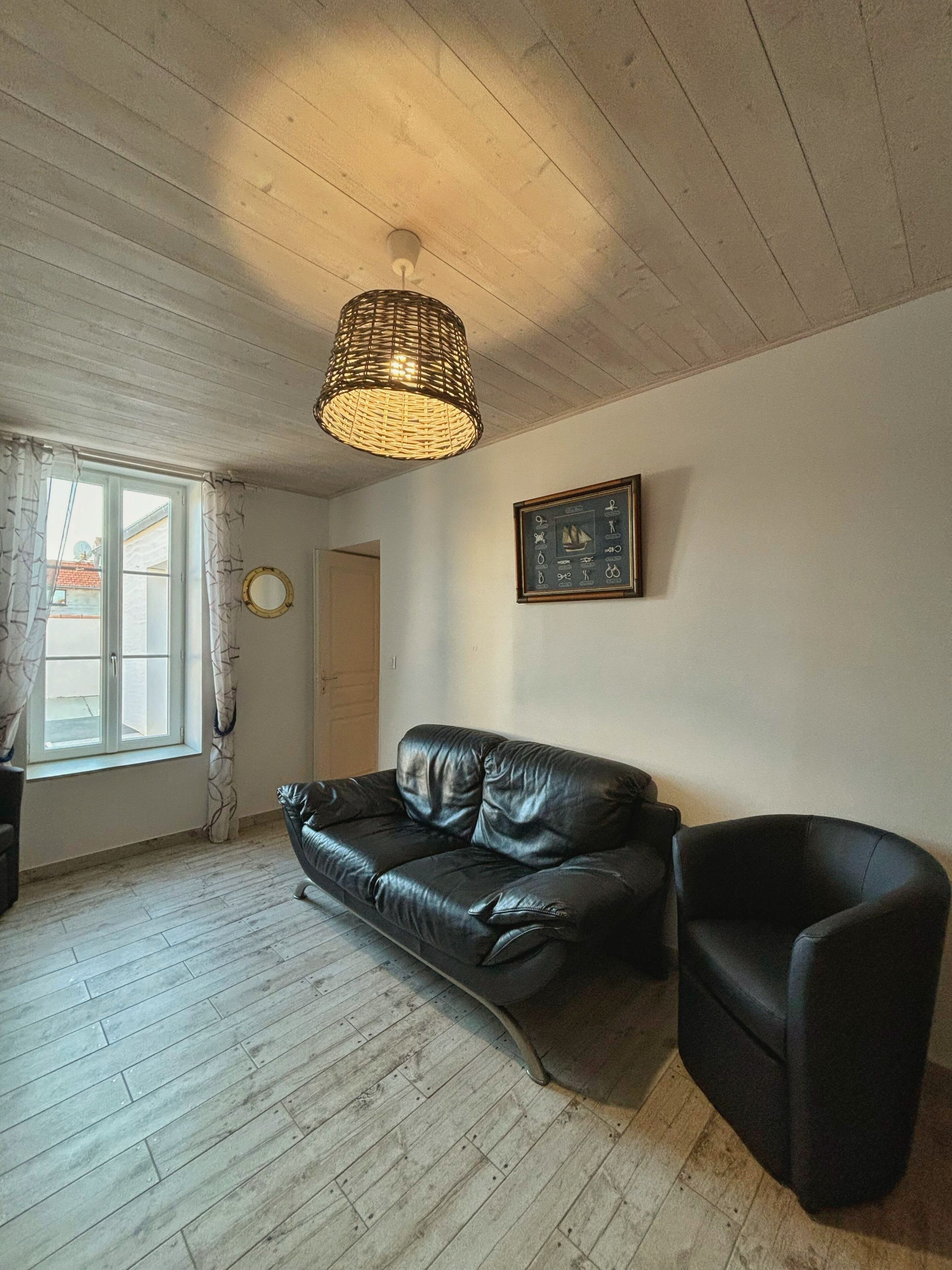 Un salon doté d’un plancher en bois, d’un canapé en cuir noir, d’un fauteuil noir, d’un luminaire suspendu en osier et d’un tableau encadré sur le mur, dont le thème est la marine.
