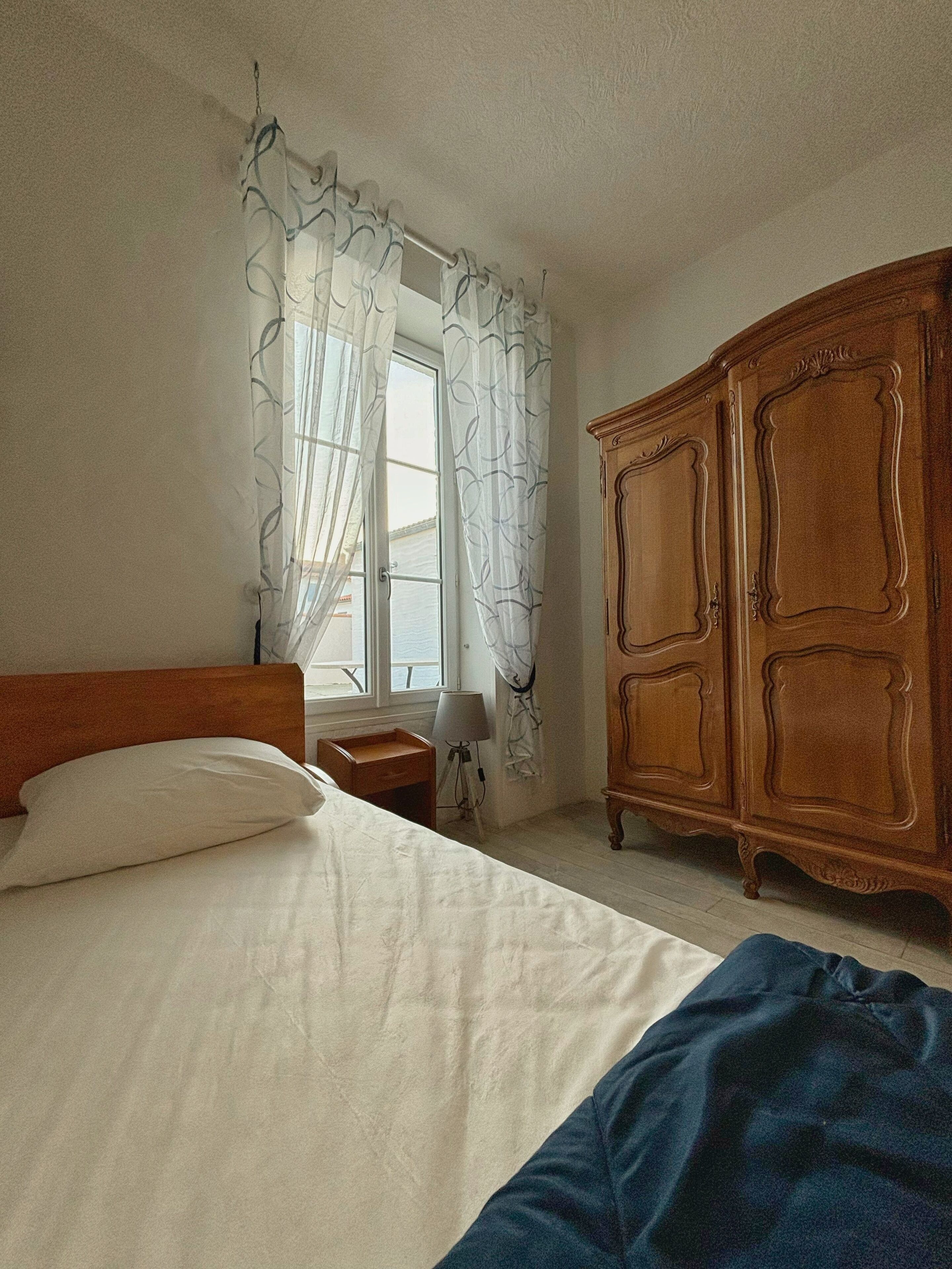 A bedroom with a bed, a wooden wardrobe, a nightstand, and a window with curtains.