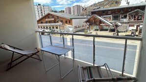 Un balcón con dos sillones reclinables y una mesa, con vista a una montaña nevada y edificios con letreros como "Restaurant Savoyare" y "Skiset".