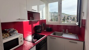 Cocina compacta con armarios blancos, encimera roja, microondas, cafetera y ventana con vistas a edificios y colinas.
