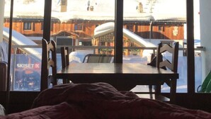 Una habitación con cama, mesa y sillas. Hay un ventanal con vistas a una montaña nevada y edificios.
