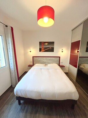 A hotel room with a bed, a red lamp, a wall-mounted picture of the Golden Gate Bridge, and a window with curtains. - CALVI new apt WIFI 3bedroom, 6 people, air-conditioned, 10mm beach (Calvi)