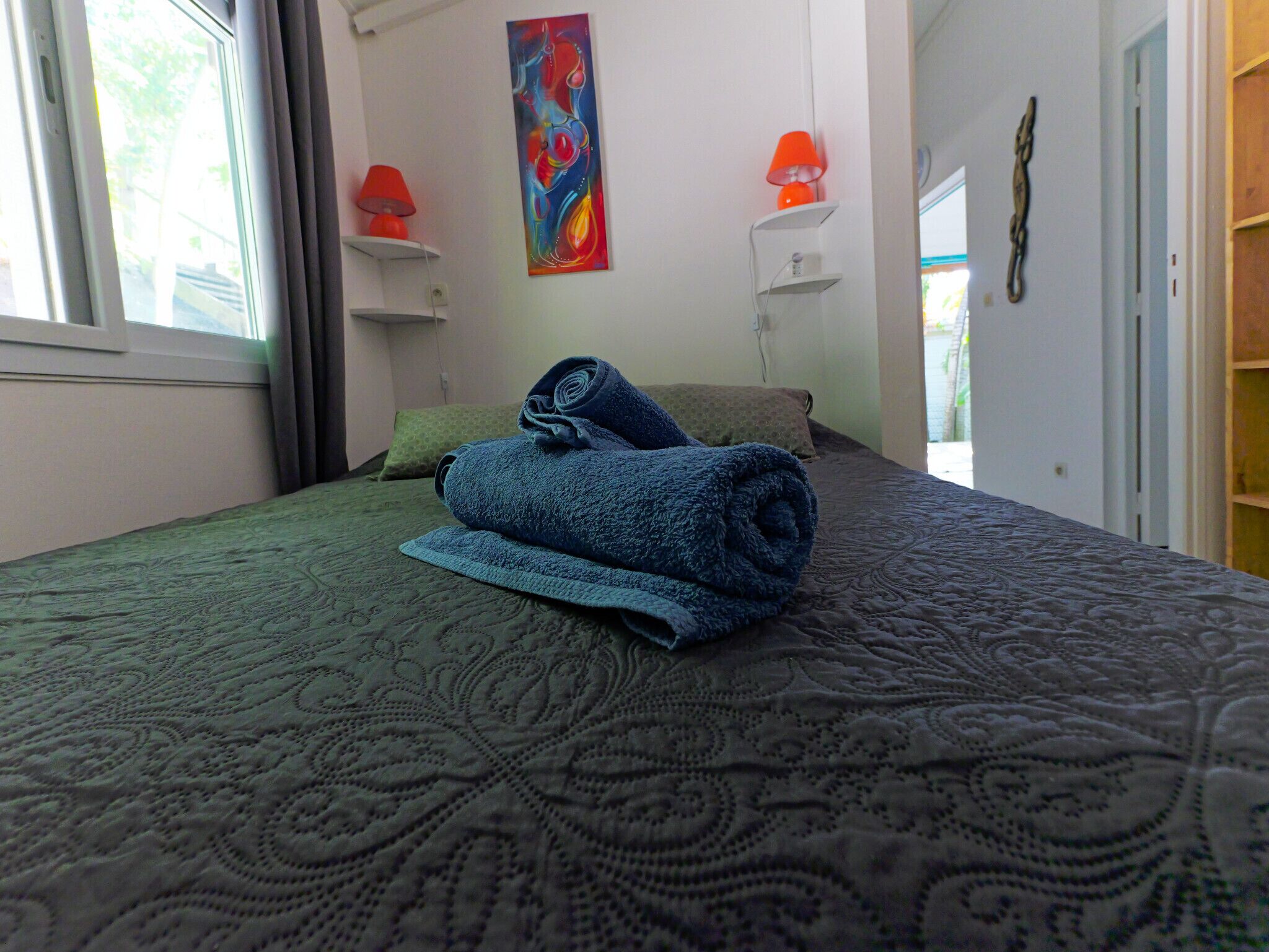 Ein ordentlich bezogenes Bett mit einem aufgerollten blauen Handtuch, einer grün gemusterten Bettdecke und zwei Kissen. An der Wand hängt ein farbenfrohes abstraktes Gemälde und zwei Wandleuchten mit orangefarbenen Lampenschirmen.