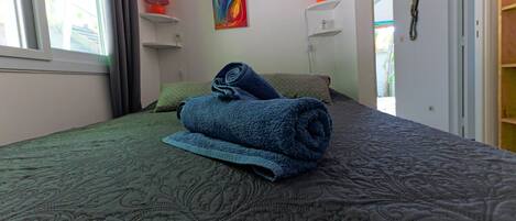 Ein ordentlich bezogenes Bett mit einem aufgerollten blauen Handtuch, einer grün gemusterten Bettdecke und zwei Kissen. An der Wand hängt ein farbenfrohes abstraktes Gemälde und zwei Wandleuchten mit orangefarbenen Lampenschirmen.
