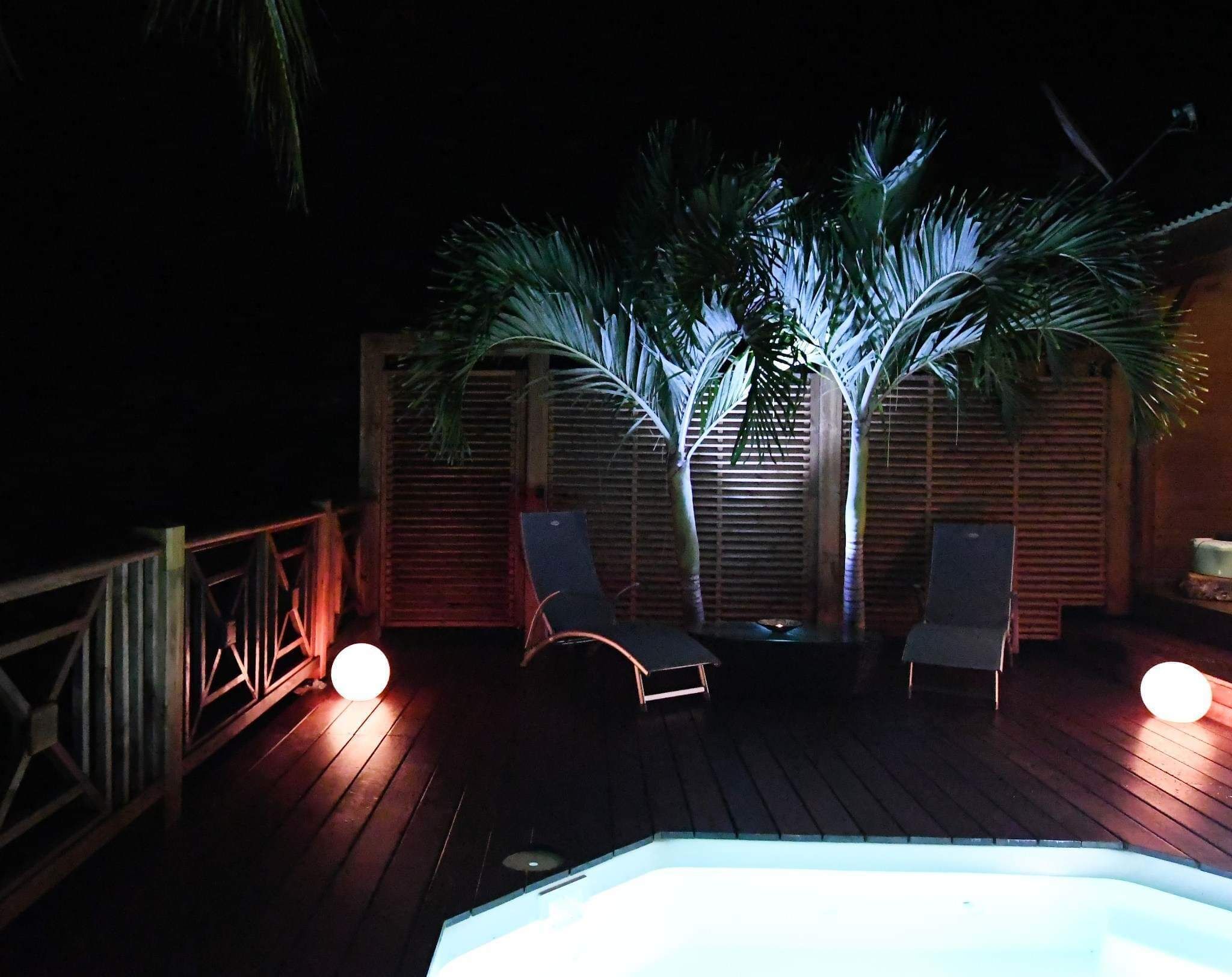 Une terrasse en bois avec deux chaises longues et un jacuzzi, entourée de palmiers et de clôtures en bois.