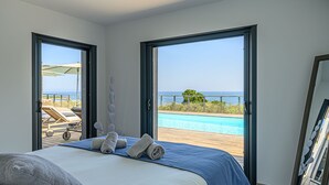 Un dormitorio con una cama, dos puertas abiertas que dan a un balcón con piscina y vistas al mar.