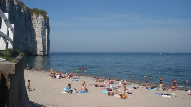 A beach with people sunbathing and swimming, cliffs in the background, and a clear sky.