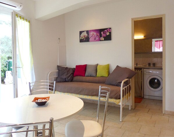 A living room with a sofa, a dining table with chairs, a washing machine, and a window with curtains. - Small house in the heart of nature, 4 km from Cargèse. (Cargèse)