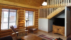 A log cabin interior with a wooden staircase, a television, and a seating area with a table and chairs. - Cabin Share Phase 2 (Angel Fire)