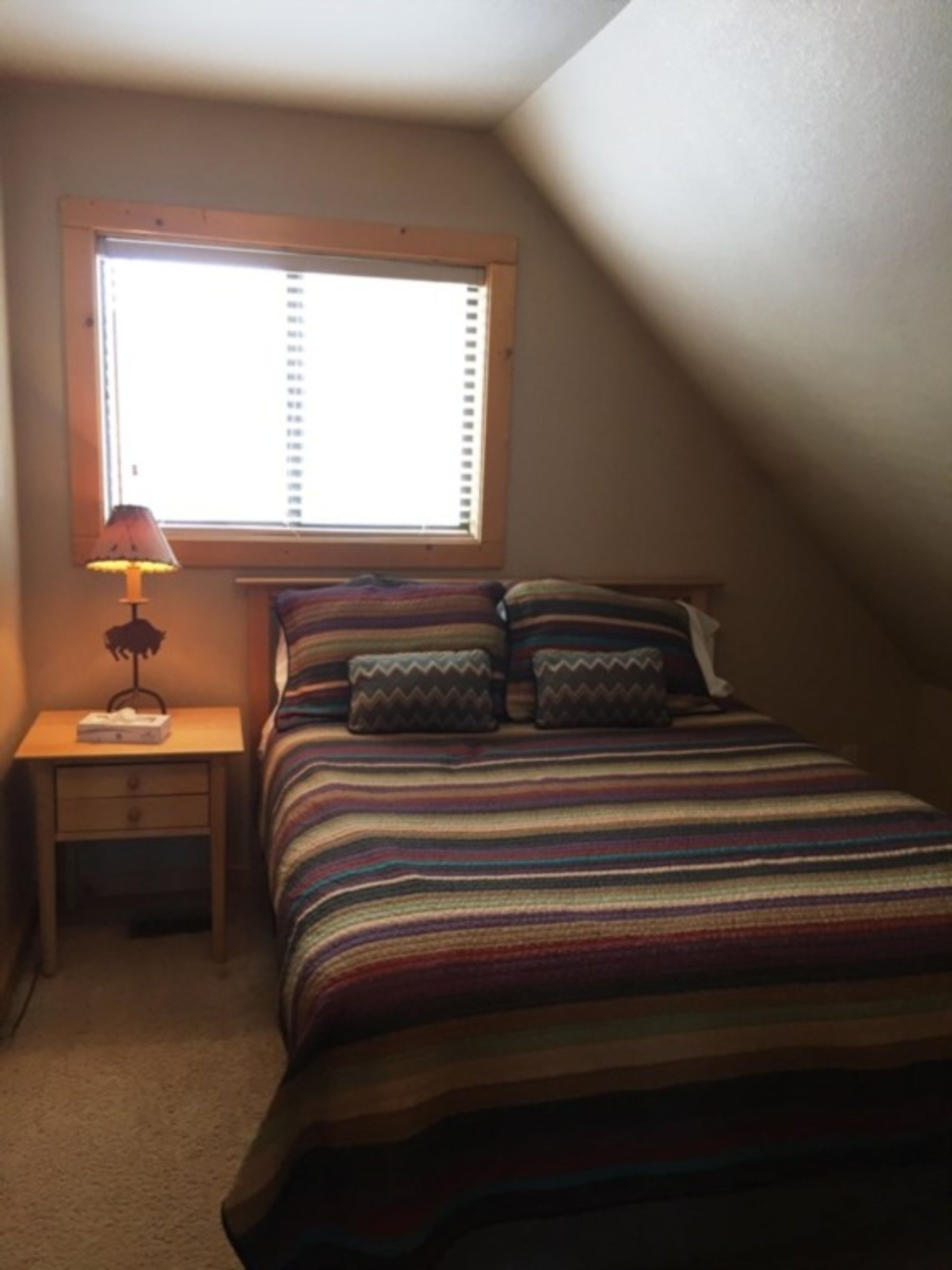 A bedroom with a bed, pillows, a nightstand, a lamp, and a window.