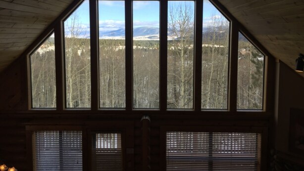 A room with a sloped ceiling and large windows offering a view of a forest and mountains. - Cabin Share Phase 2 (Angel Fire)