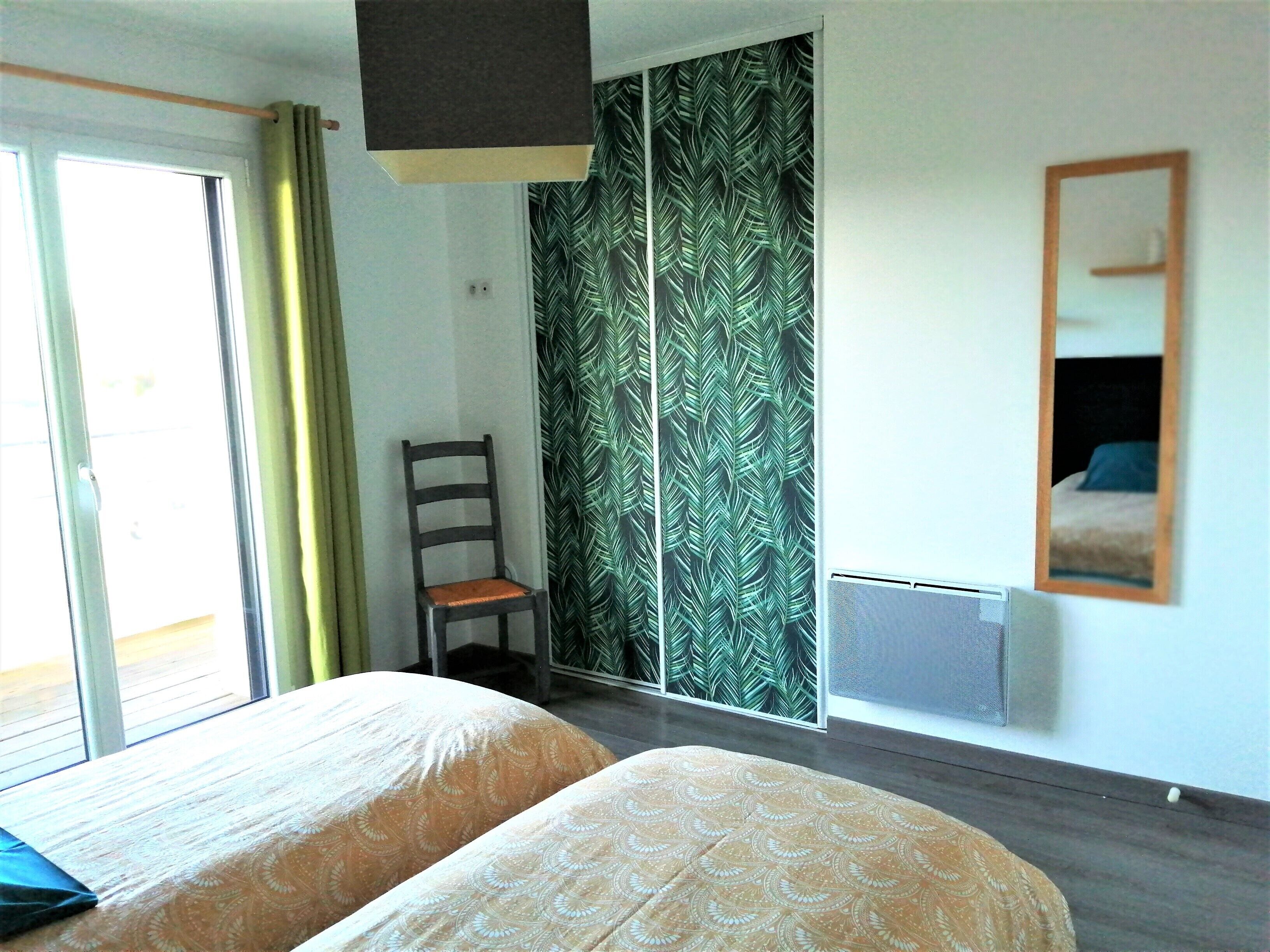 Habitación de hotel con dos camas, una silla, un ventanal con cortinas, una puerta corrediza con diseño de hojas y un espejo en la pared.
