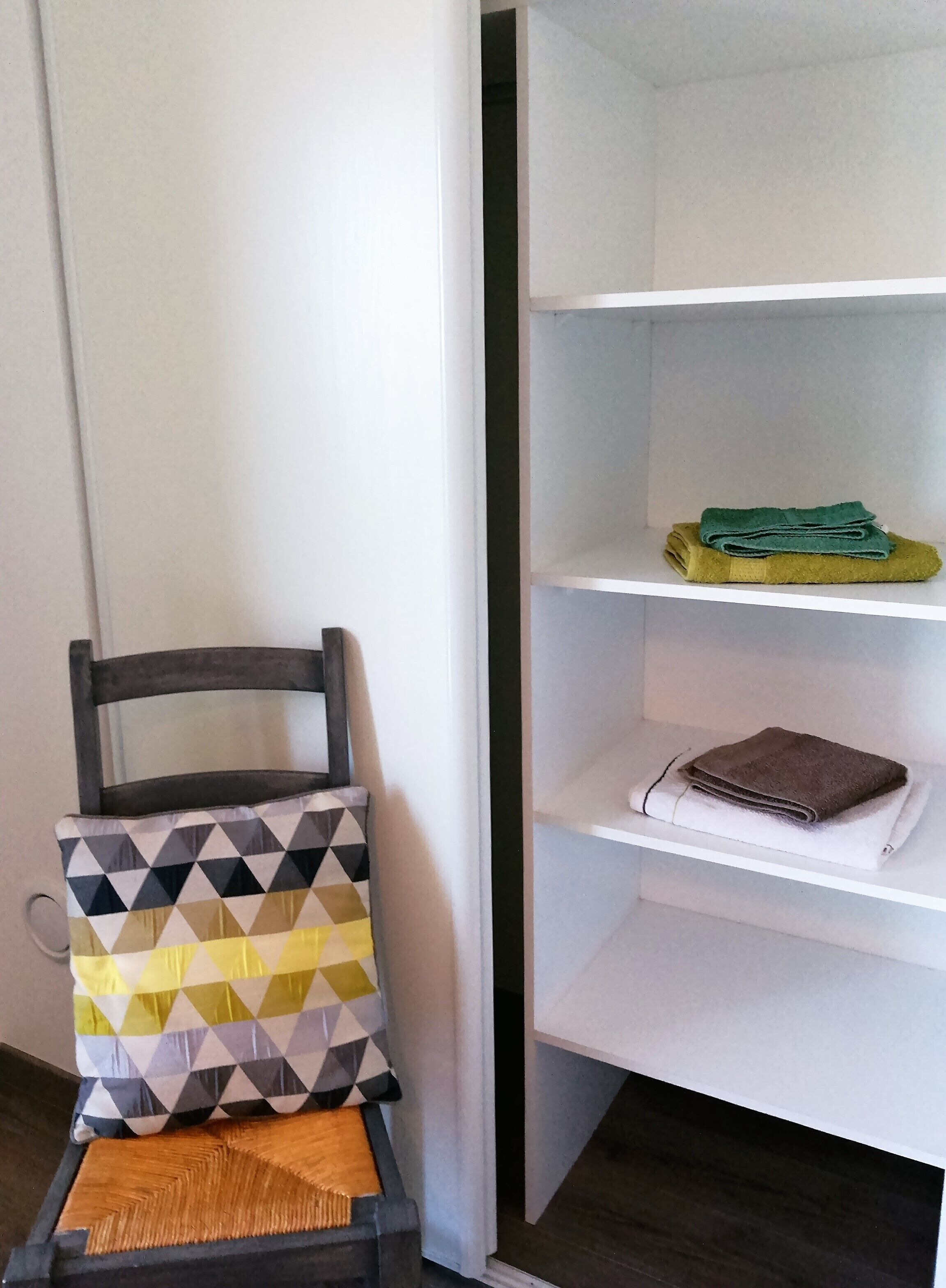 Une pièce avec une chaise, un coussin à motifs et une étagère blanche où sont pliées des serviettes.