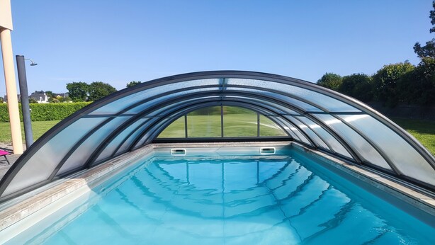 A swimming pool with a glass enclosure and a view of a green lawn and houses in the background. - Modern villa in Les Sables d'Or with heated pool (Plurien)