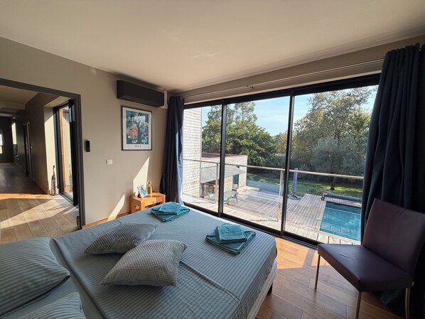 A bedroom with a large window, a bed with pillows, a chair, and a small table with a picture frame.