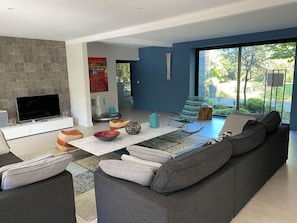 Uma sala de estar moderna com um sofá cinzento, uma televisão de tela plana, uma lareira e uma grande janela com vista para o verde.