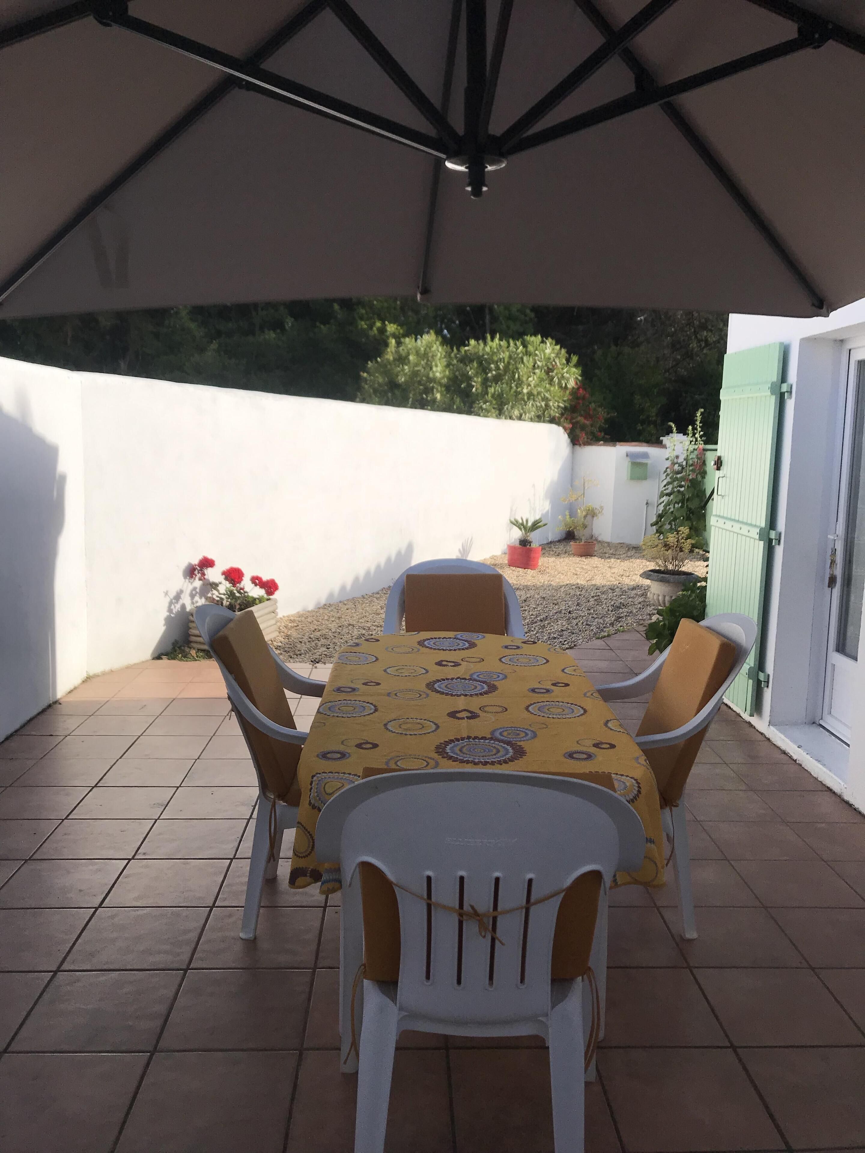 A covered patio area with a table and chairs, surrounded by a white wall and potted plants.