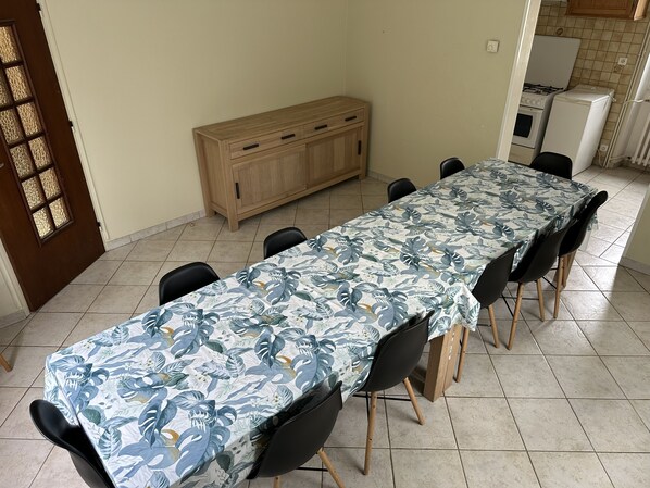 A dining area with a long table covered by a tropical leaf tablecloth, surrounded by black chairs with wooden legs. A wooden cabinet is against the wall, and a kitchen area with a stove and refrigerator is visible in the background.