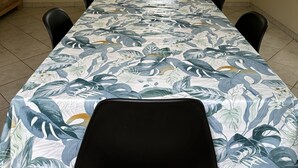 Ein Esszimmer mit einem langen Tisch, der mit einer Tischdecke aus tropischen Blättern bedeckt ist, schwarzen Stühlen, einem Kronleuchter und Blick auf eine Küche durch eine Tür.