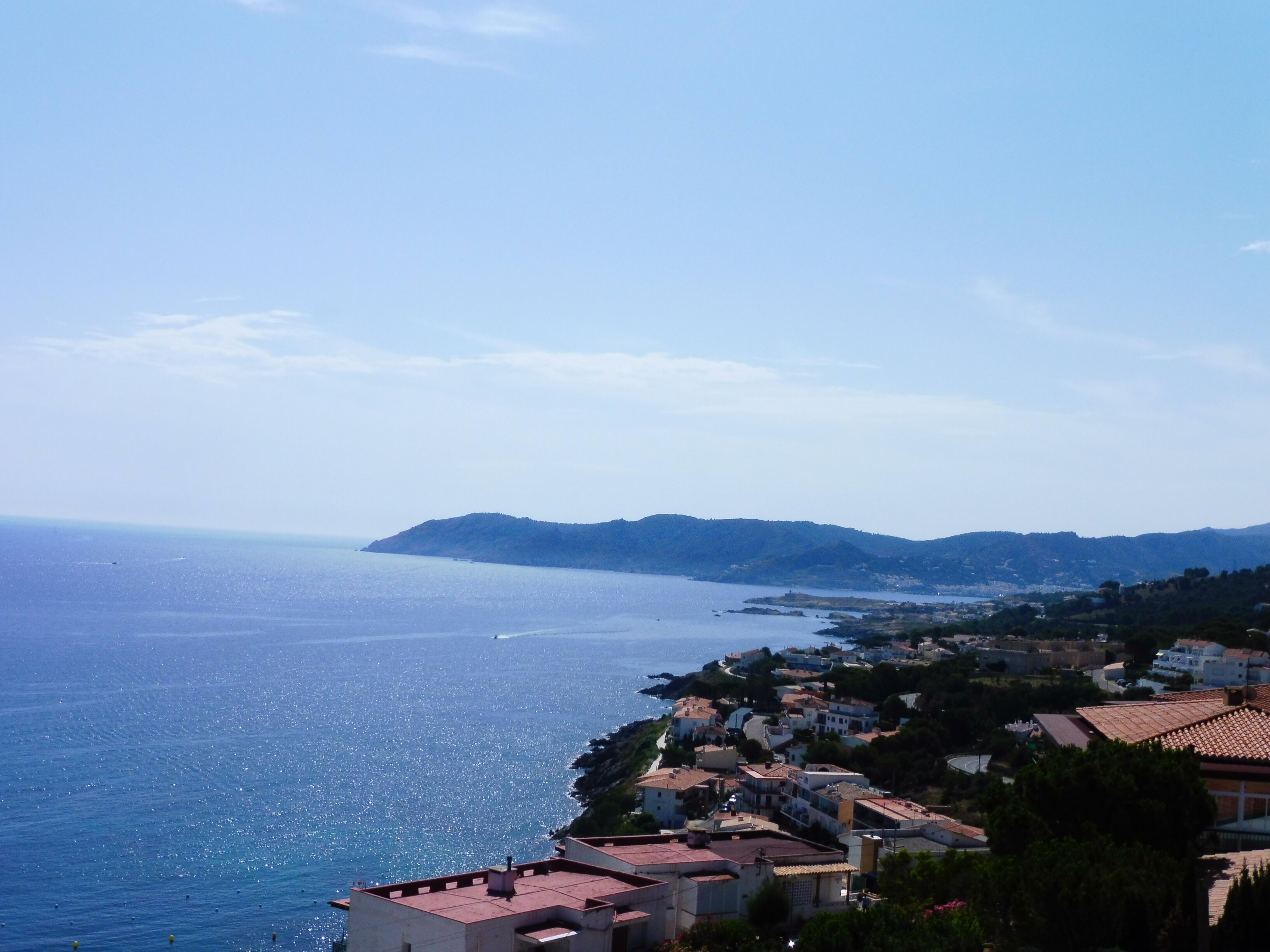 A coastal town with buildings along the sea, a clear sky, and distant hills.